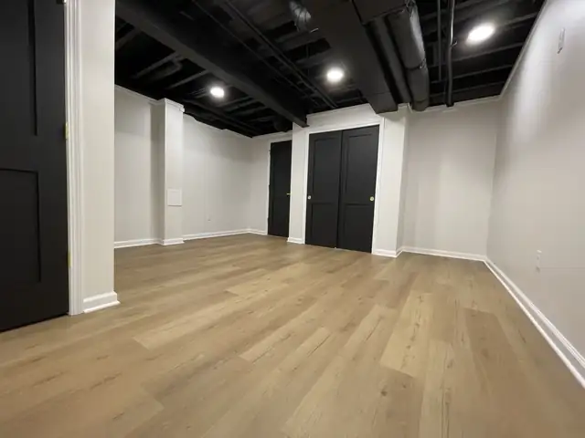 Finished basement with black painted open ceiling, recessed lights, black doors and light wood-look vinyl floor