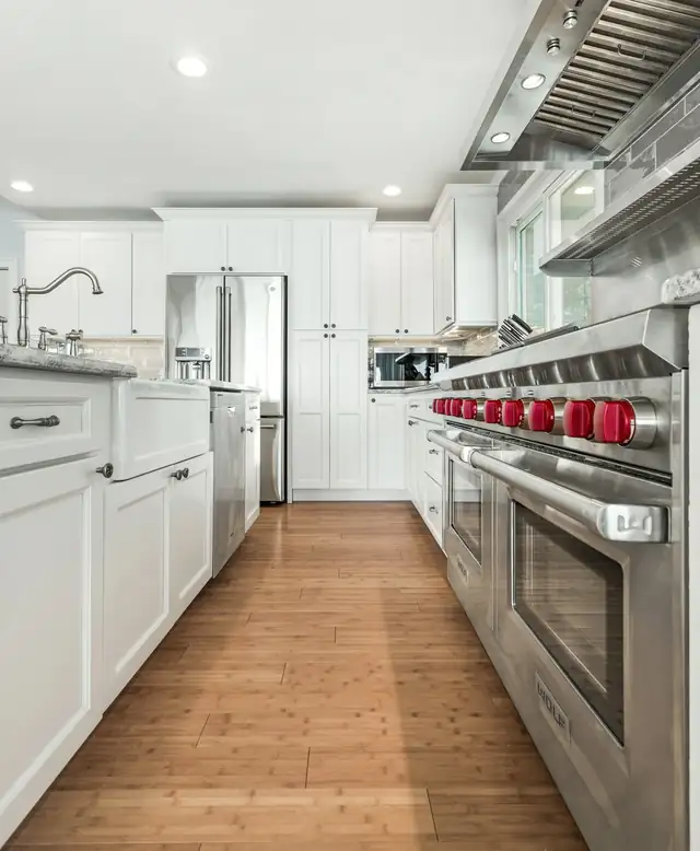 White galley kitchen by Rohrer For Construction with a farmhouse sink, a stainless range with red knobs and wood-look flooring